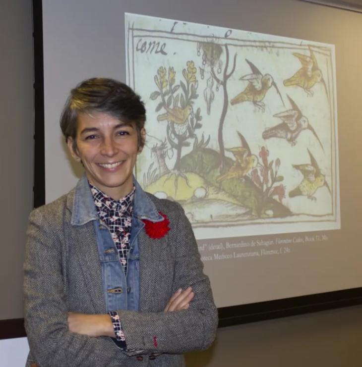 Professor Montero has short hair and a bright smile, she is of Mexican heritage and stands in front of a power point projection displaying a collaboratively written document between Mexican and the Spanish (Humming bird painting included)