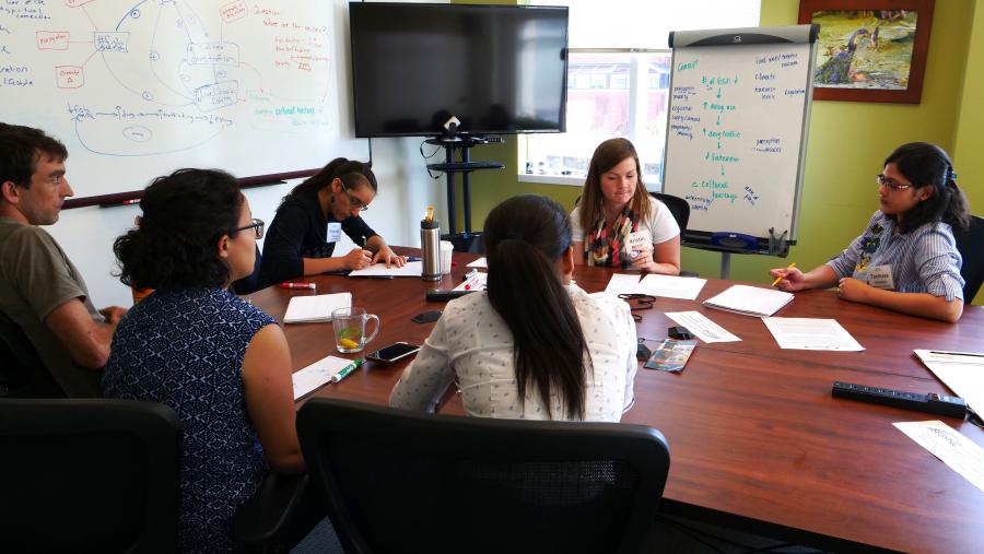 A research team gathered around a table with writing on a whiteboard in the background