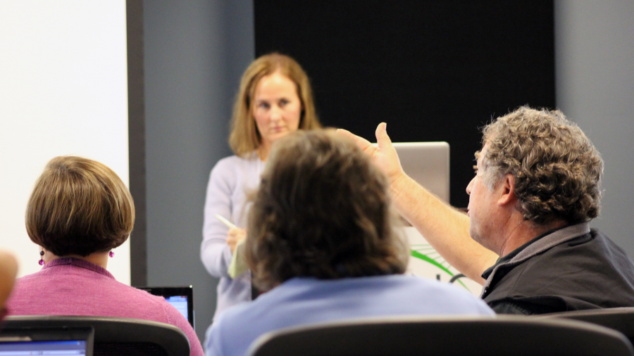 Three people seated with their backs to the camera with one woman standing facing them as they engage in a discussion