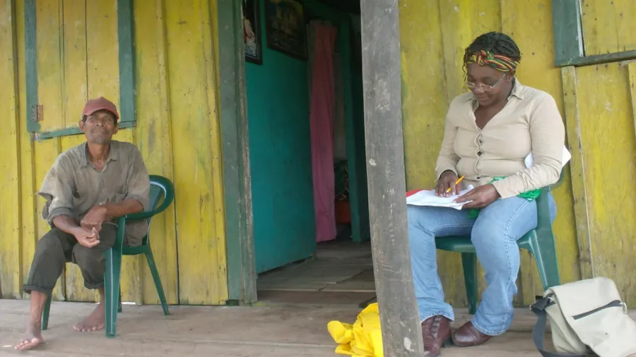 A man and woman seated on the porch of a yellow house with the woman writing down notes while interviewing the man