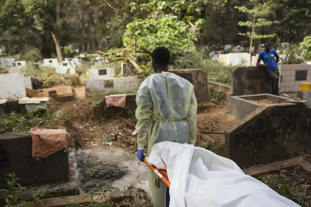 A worker carrying a stretcher with a body covered by a white cloth into a graveyard