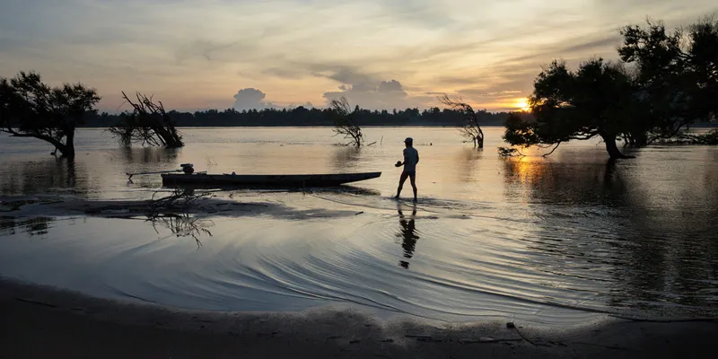 The silhouette of a man walking in the shallow water of a lake toward a long, narrow boat