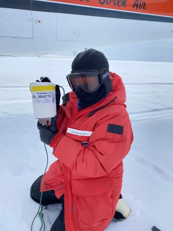 A photo of Kathrine Udell kneeling on the ground, wearing a red coat and face mask, posing with equipment in Antarctica