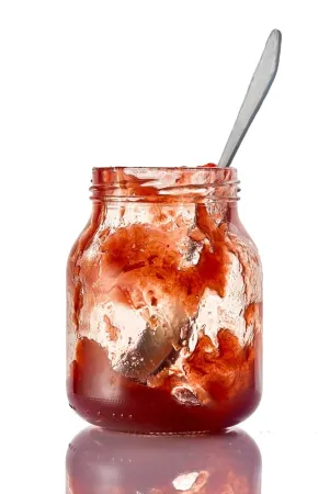 A nearly empty jar of jam with a spoon inside in front of a white background