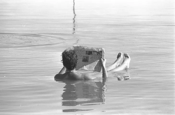 A black-and-white photo of a person reading a newspaper while floating in the Dead Sea