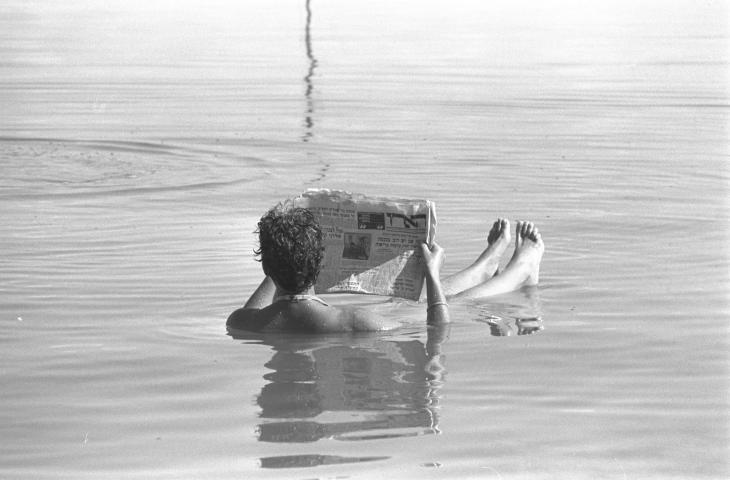 A black-and-white photo of a person reading a newspaper while floating in the Dead Sea