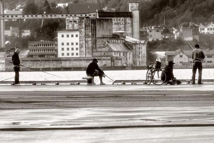 A black-and-white photo showing four men fishing from a dock in an urban setting