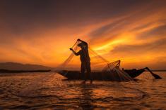 A man in silhouette against a sunset standing in a small fishing boat bringing in a net