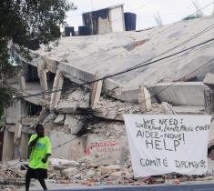A man standing in front of a collapsed building with a banner on the rubble saying "Help"